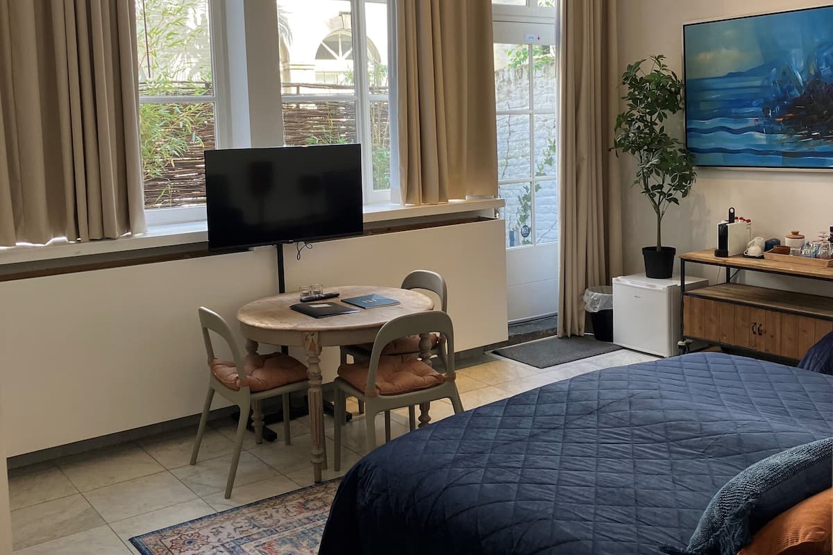 A bedroom with a bed, a desk with a chair, a television, a plant, and a painting on the wall.