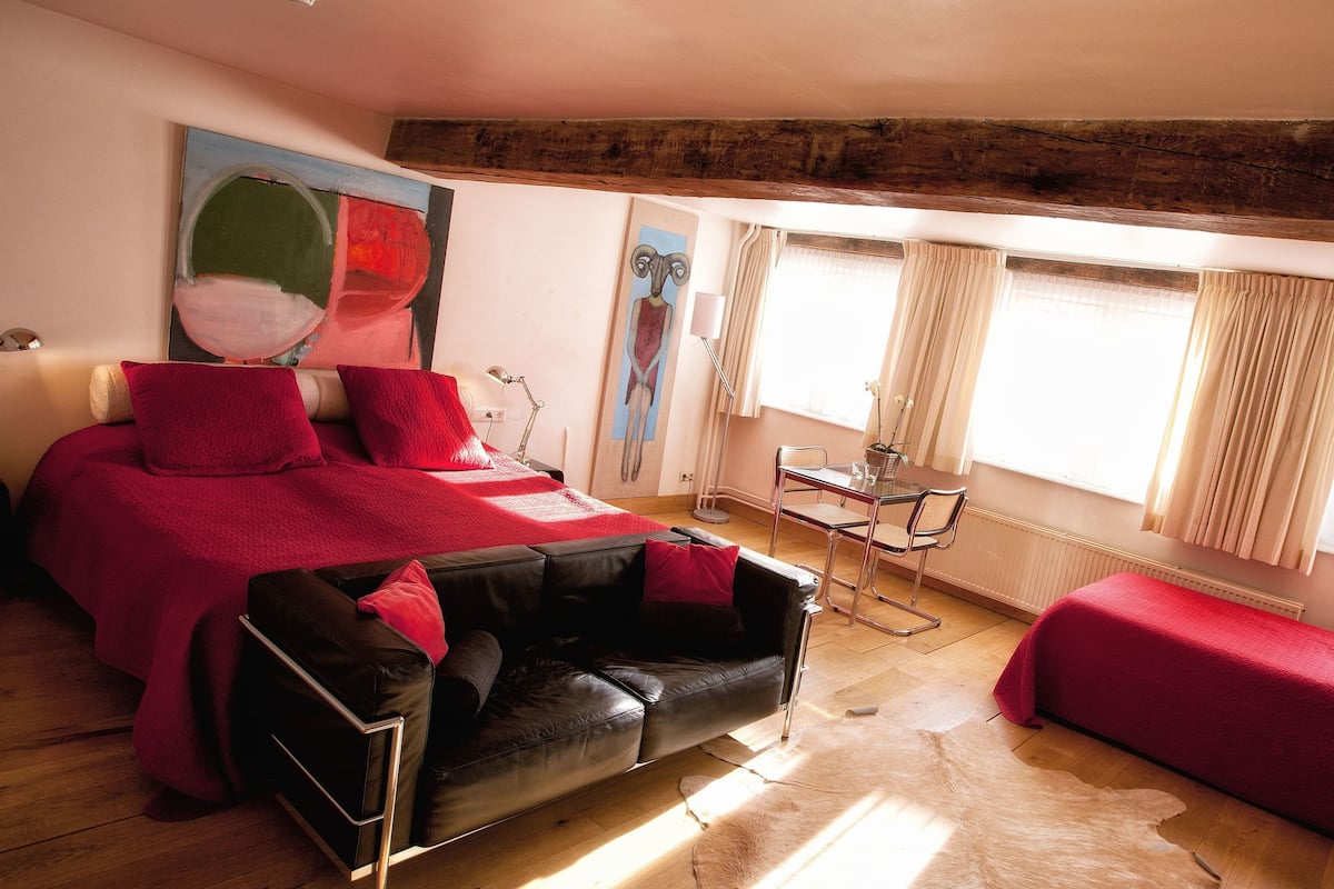 A modern bedroom with a bed, a black leather sofa, a red sofa, a dining table with chairs, and a large window with curtains.