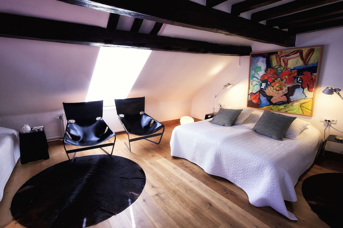A cozy loft bedroom with a sloped ceiling, wooden beams, a large window, a bed with pillows, two black chairs, and a painting on the wall.