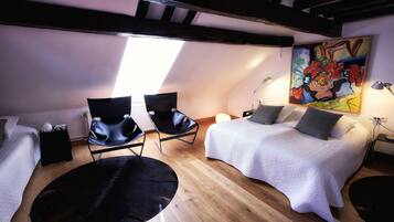 Une chambre à coucher confortable, aménagée dans un grenier avec un plafond en pente, des poutres en bois, une grande fenêtre, un lit avec des oreillers, deux chaises noires et un tableau accroché au mur.