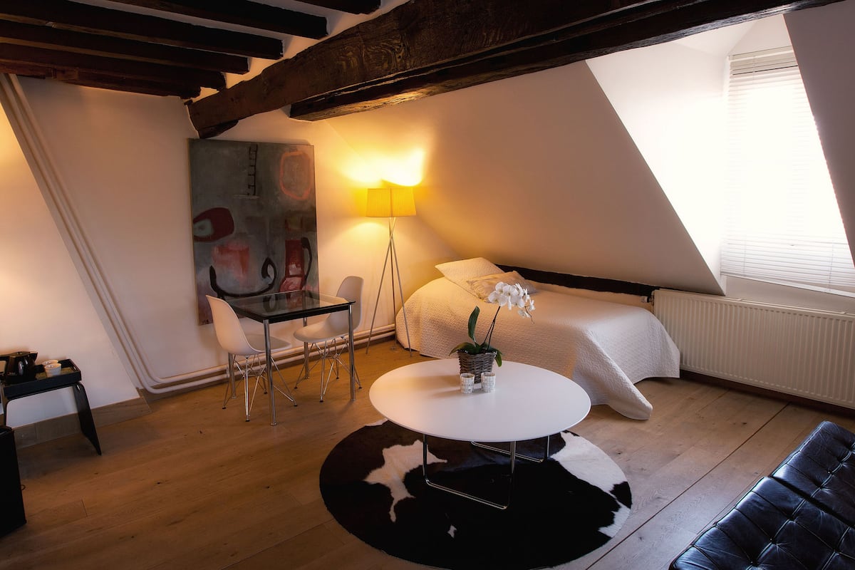 A cozy, well-lit room with a bed, a round table with chairs, a lamp, and a painting on the wall.
