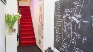 Un pasillo con alfombra roja que conduce a una escalera, una pizarra con un mapa y un florero con flores blancas.
