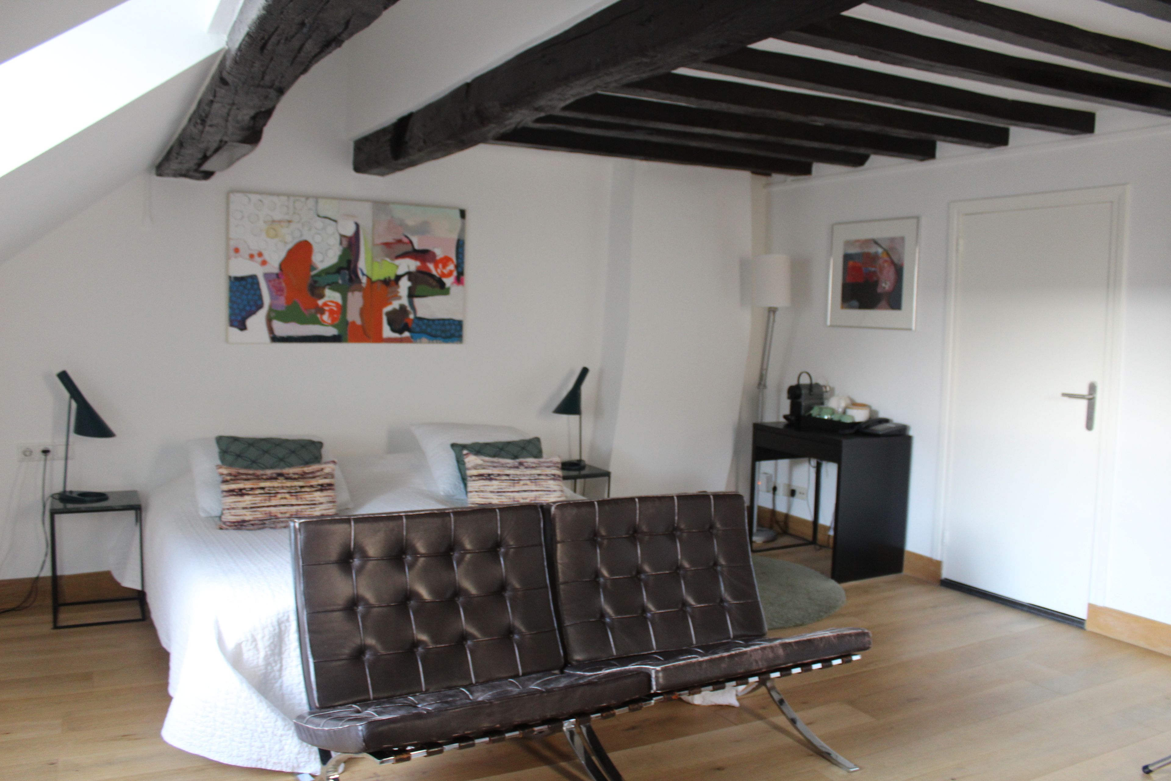 Ein Zimmer mit einem Bett, einer braunen Polsterbank, einem Gemälde an der Wand und einer Tür.