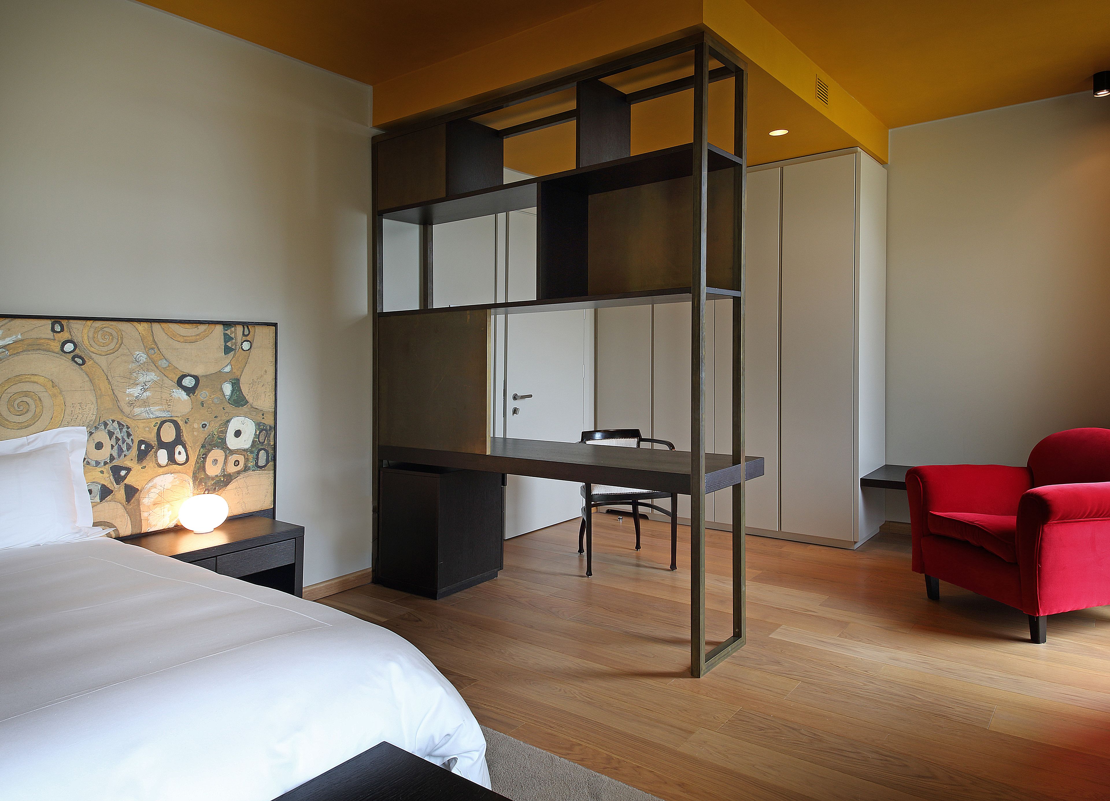 A modern hotel room with a large bed, a desk, a red chair, and a unique wall art piece.