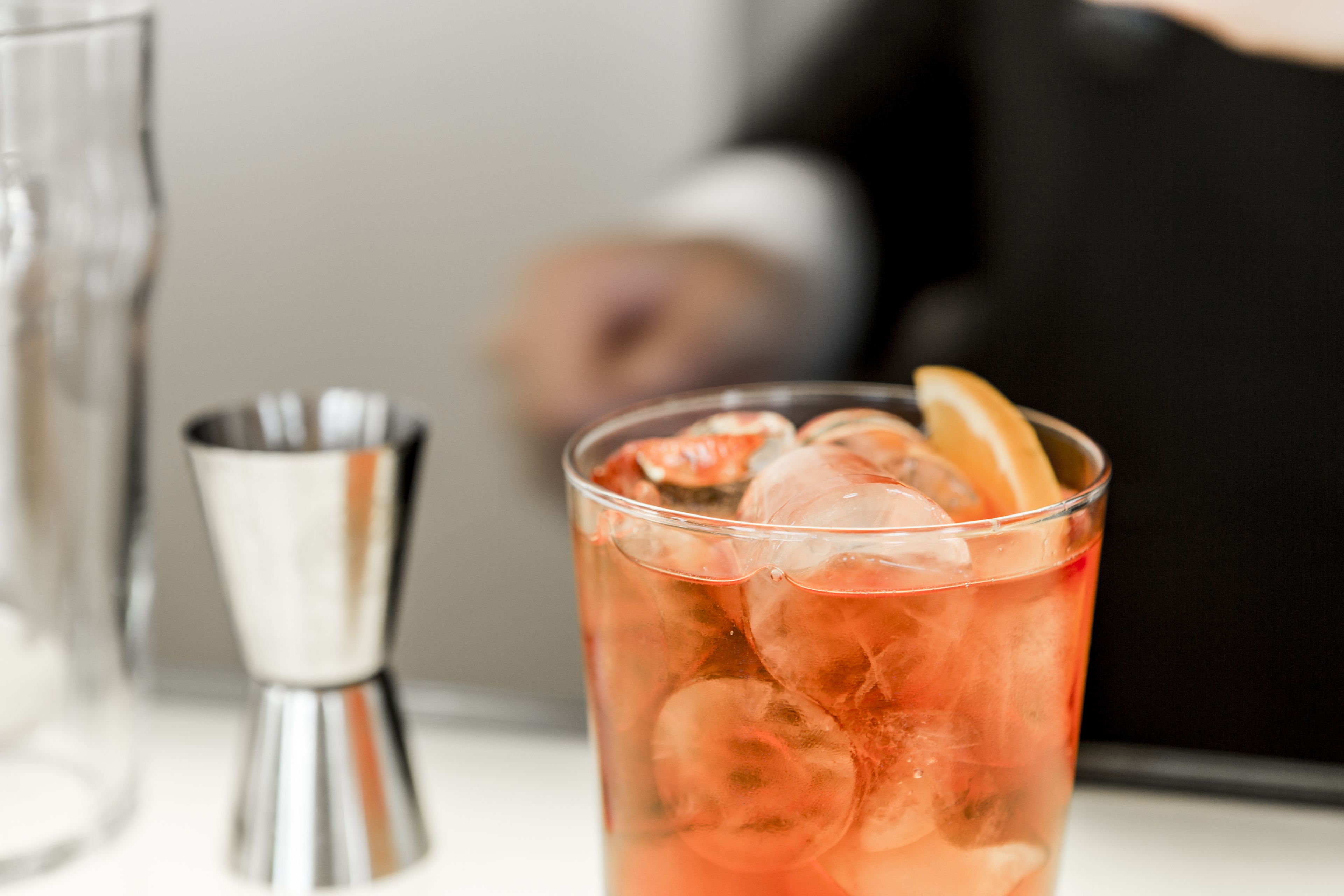 Et glas med iskold drik, isterninger og et skive appelsin, en cocktailshaker og en uskarpt baggrund.