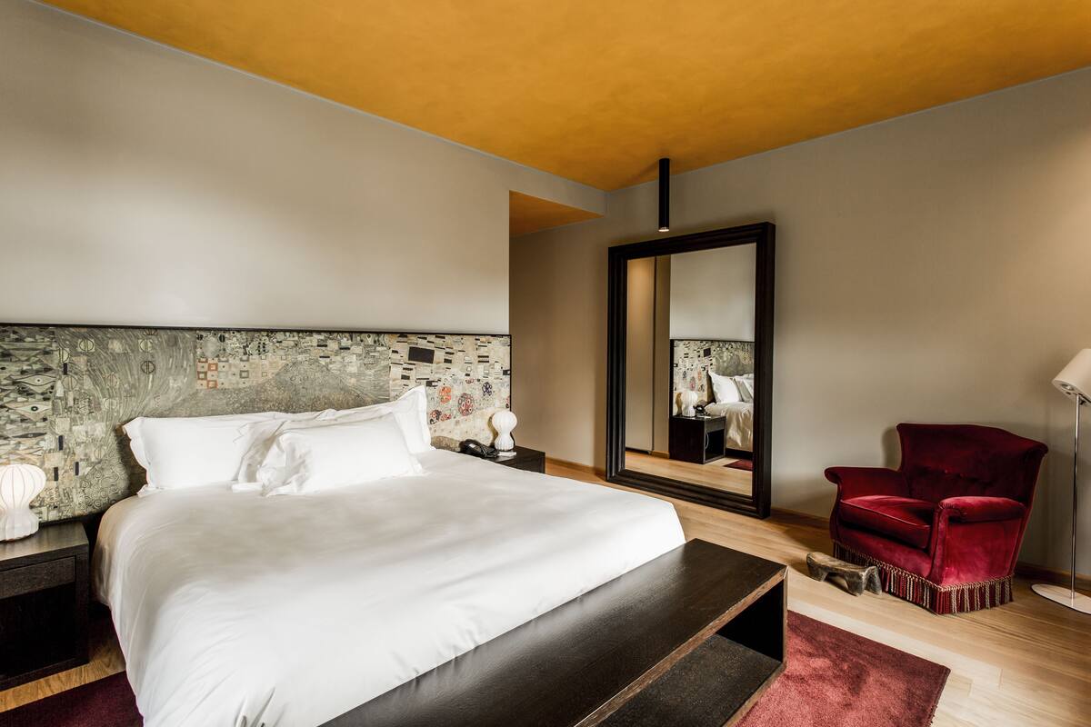 A modern hotel room with a large bed, a red armchair, a mirror, and a wooden nightstand.