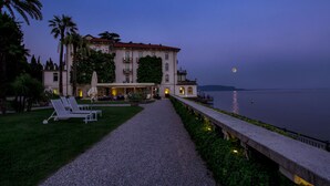 A waterfront hotel with a gravel pathway leading to a seating area with lounge chairs. - Hotel Bella Riva (Gardone Riviera)
