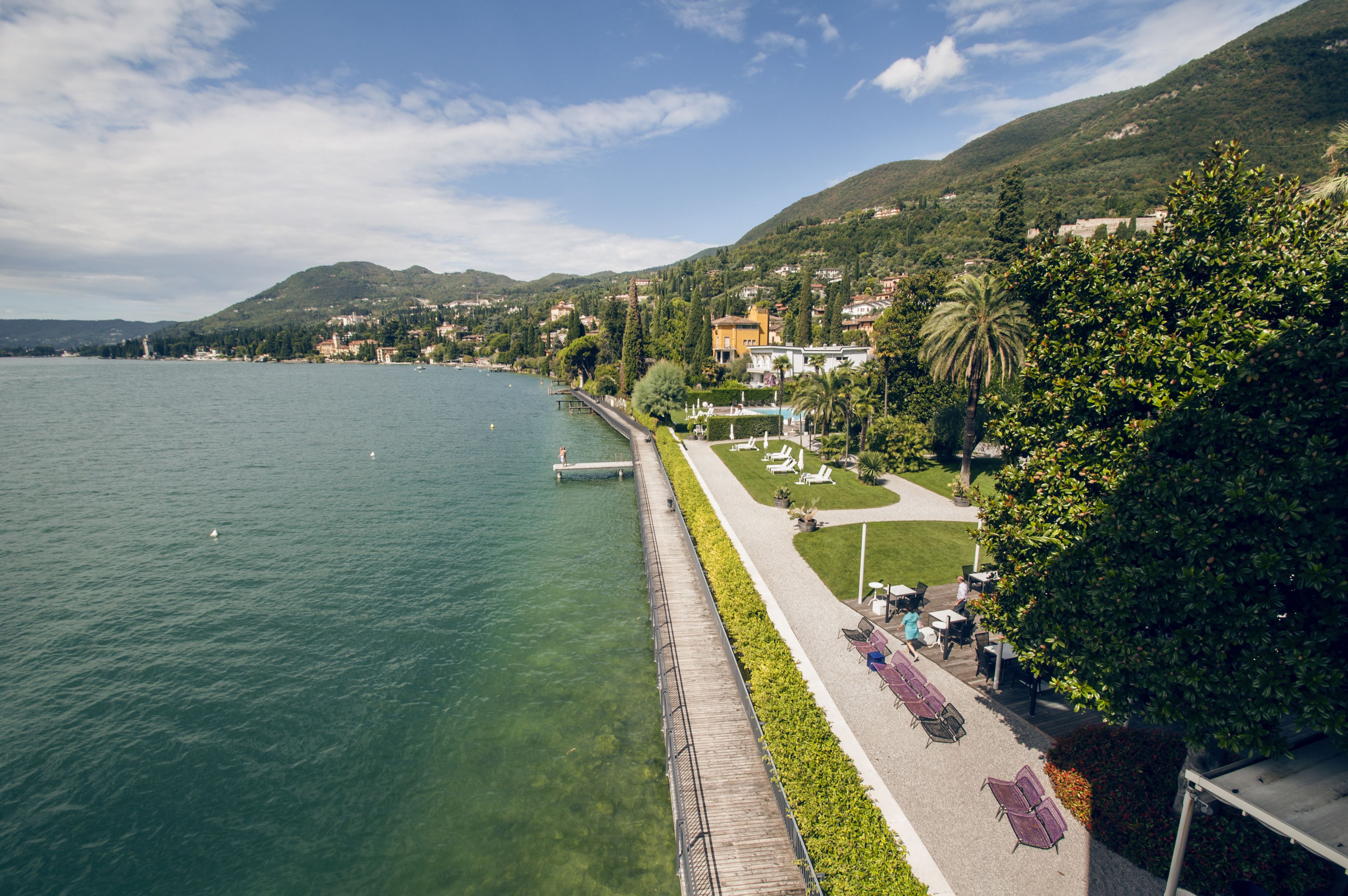 Eine Uferpromenade mit Holzsteg, Sitzbereichen und Blick auf den See und die umliegenden Hügel.