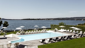 A poolside area with lounge chairs and umbrellas, a wooden deck, and a view of the water. - Hotel Bella Riva (Gardone Riviera)