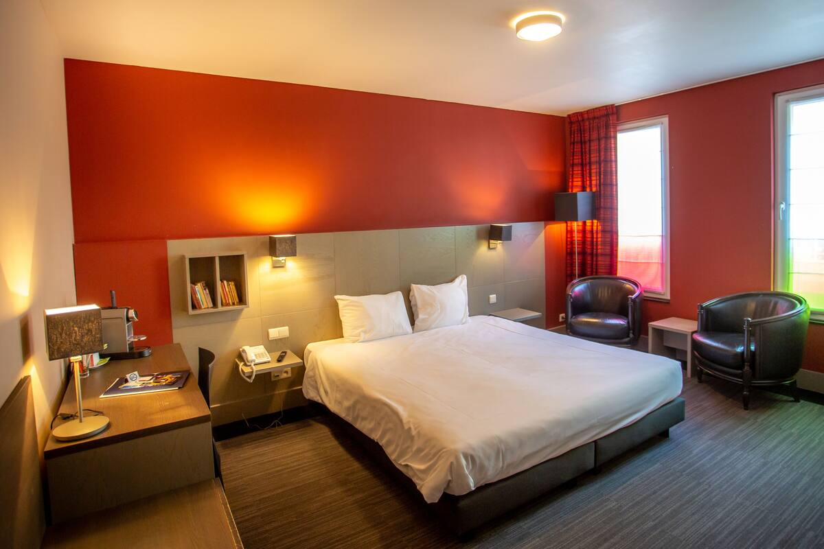 A hotel room with a large bed, bedside tables, a desk, a chair, and a window with curtains.
