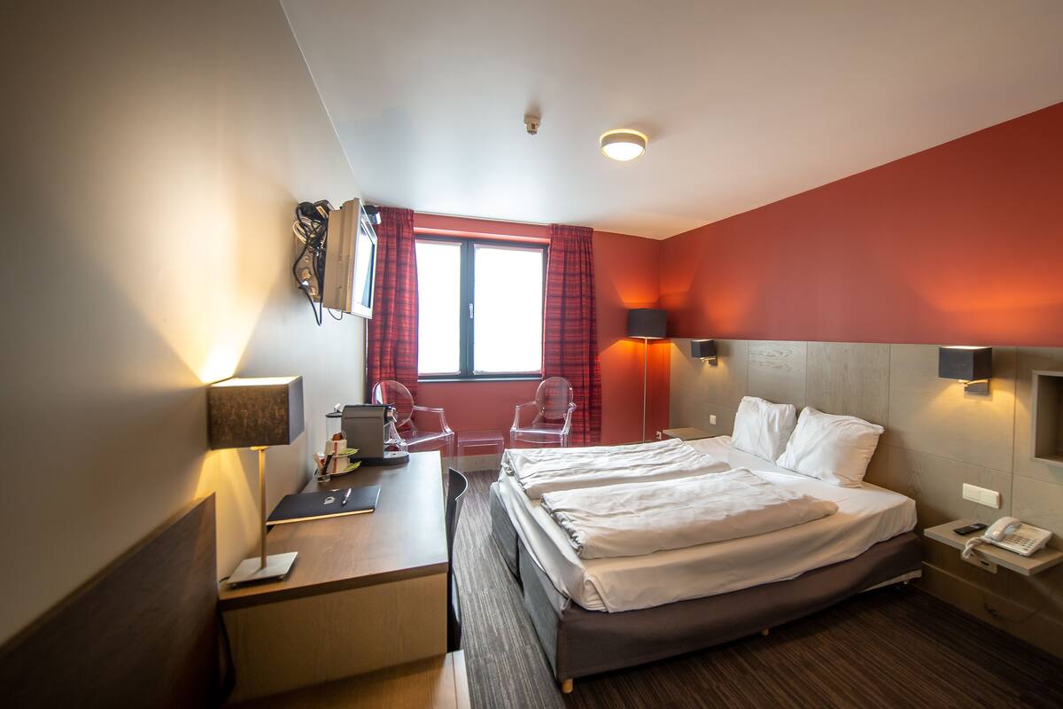 A hotel room with a bed, desk, chair, and a small shelf with books.