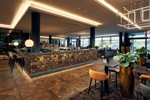 A modern bar area with a geometric patterned floor, bar stools, and a central counter with a mosaic tile design. - Van der Valk Hotel Nivelles - Sud (Nivelles)
