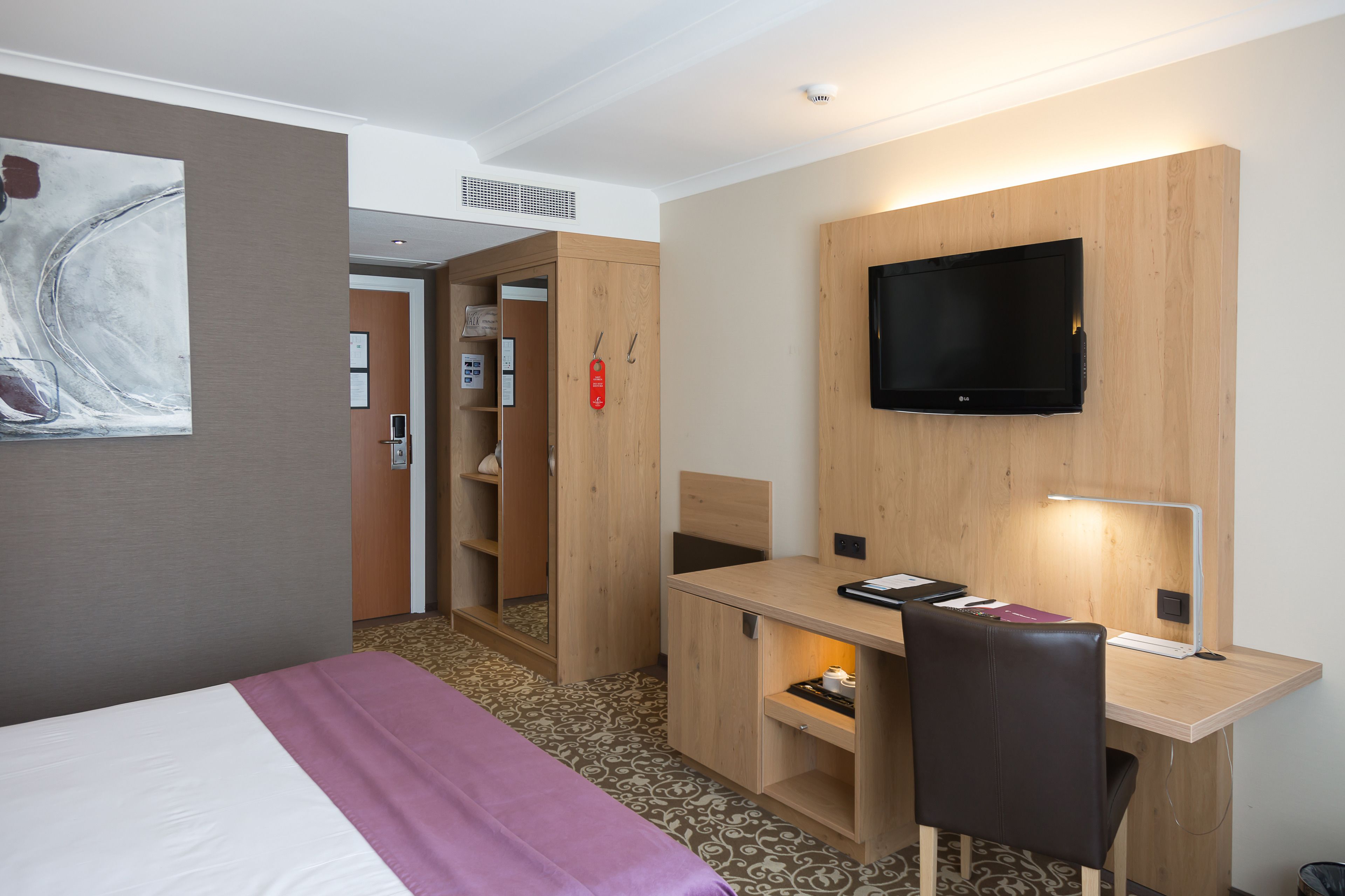 A hotel room with a bed, a desk with a chair, a television mounted on the wall, and a small shelf with items.