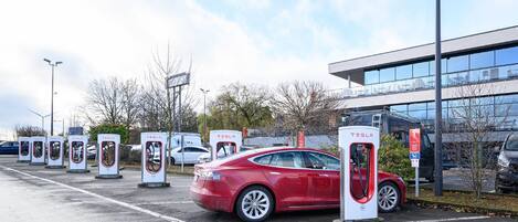 Una estación de carga de Tesla en un estacionamiento, con un edificio moderno al fondo.