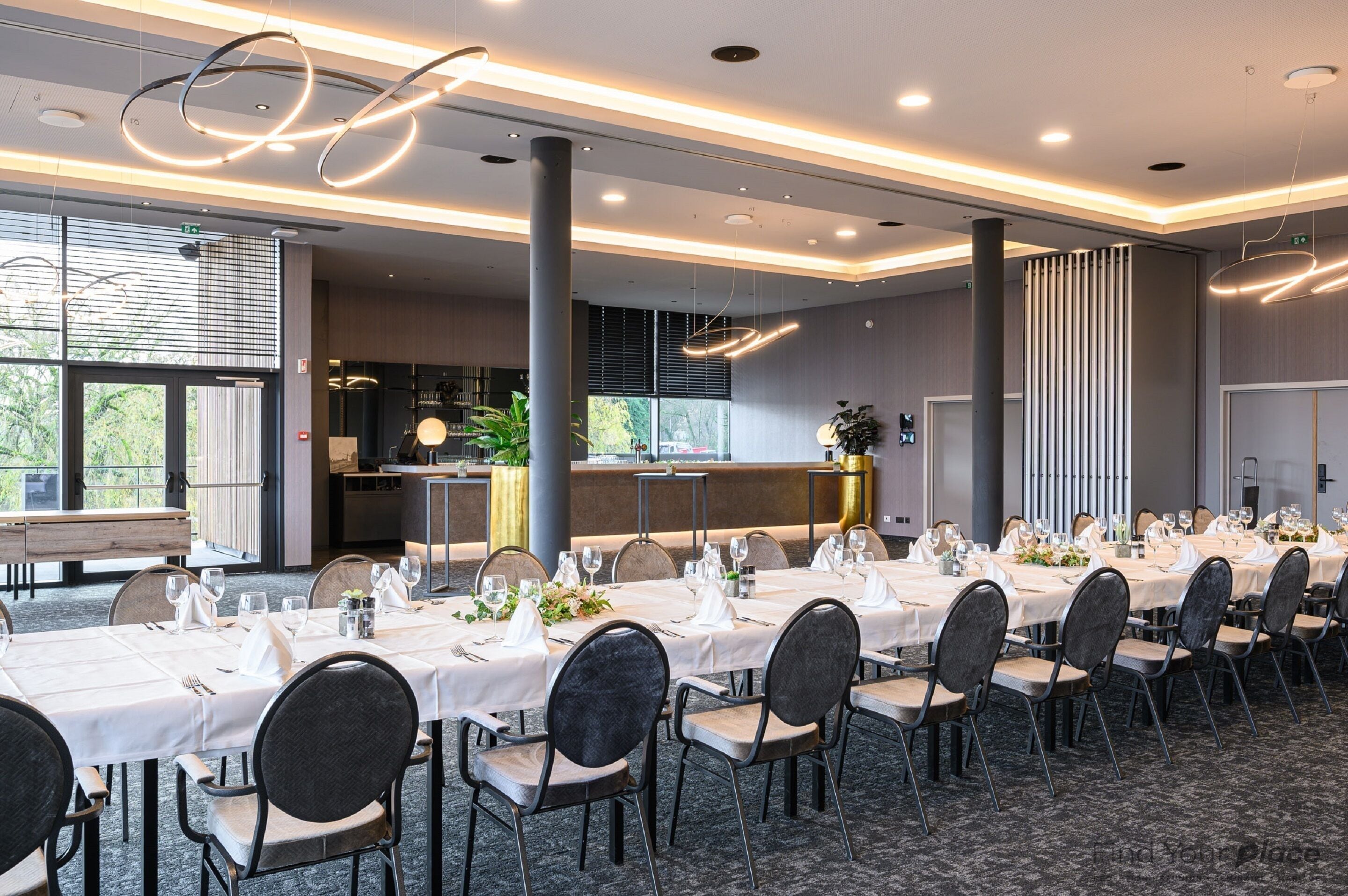 A conference room with long tables set for a formal event, featuring modern lighting and a view of the outdoors.