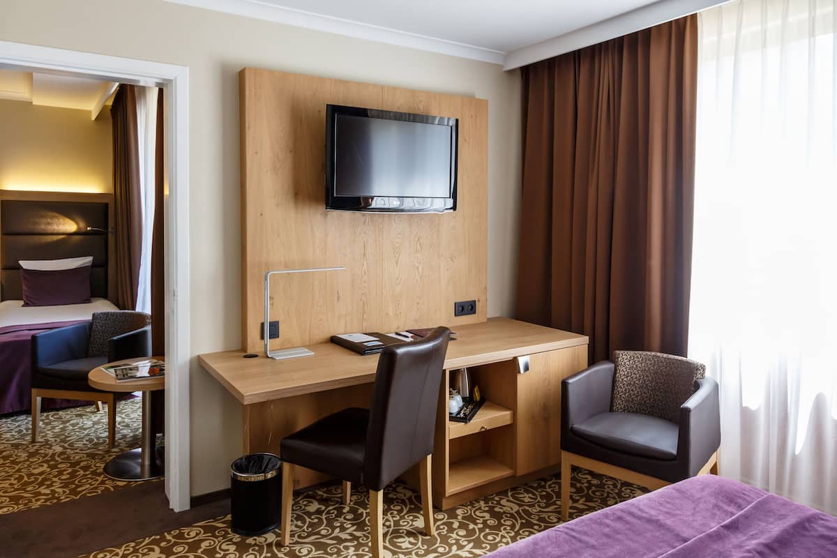 Una habitación de hotel con un escritorio de madera, una silla, un televisor montado en la pared, una cama con colcha morada y una alfombra estampada.
