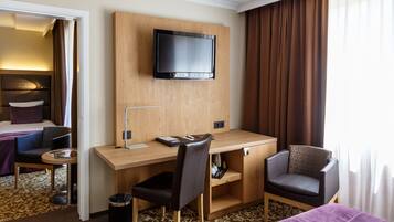 Een hotelkamer met een houten bureau, een stoel, een aan de muur bevestigde televisie, een bed met een paars sprei en een kamerbreed tapijt met dessin.