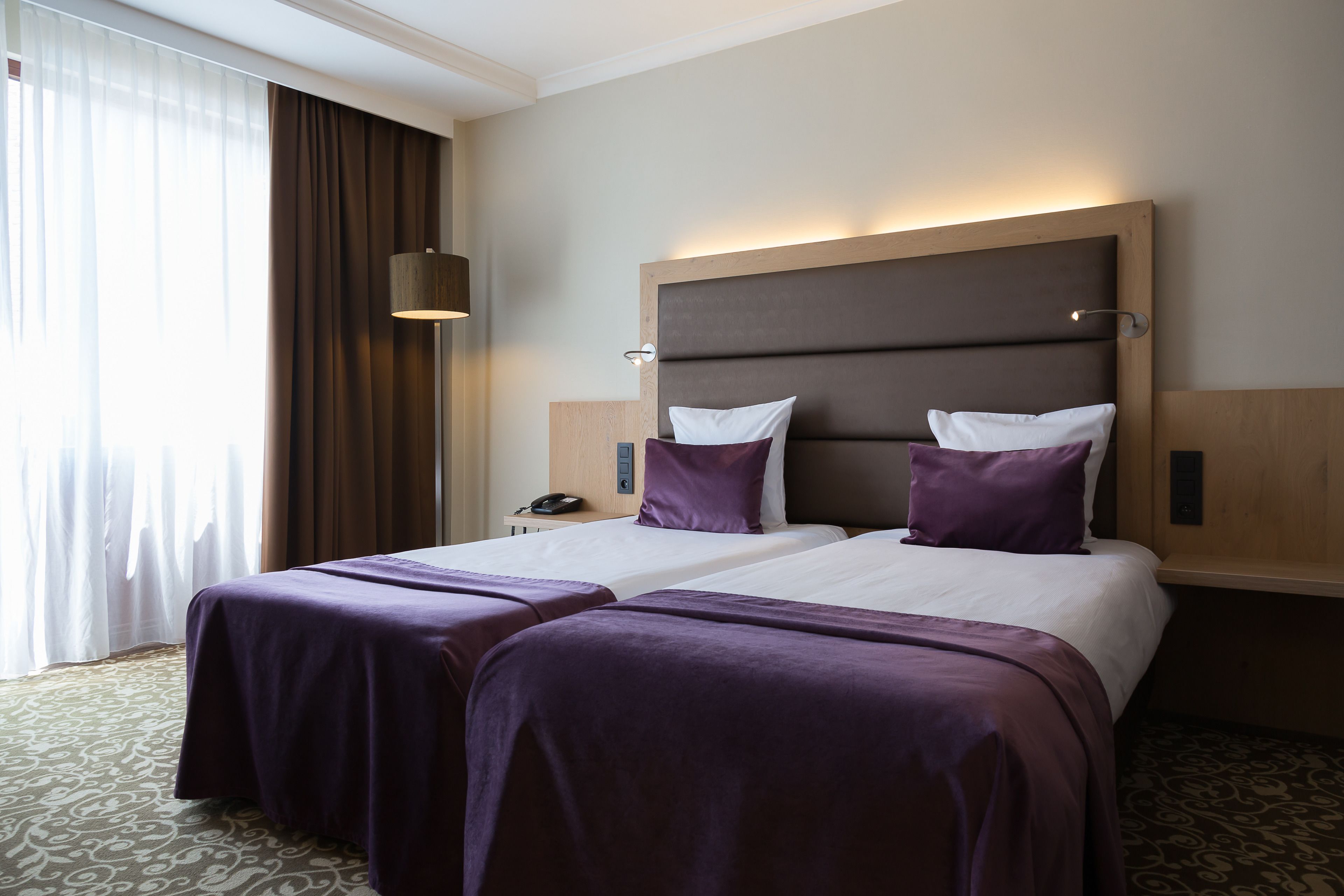 A hotel room with two beds, a large window with curtains, a bedside table with a lamp, and a phone.