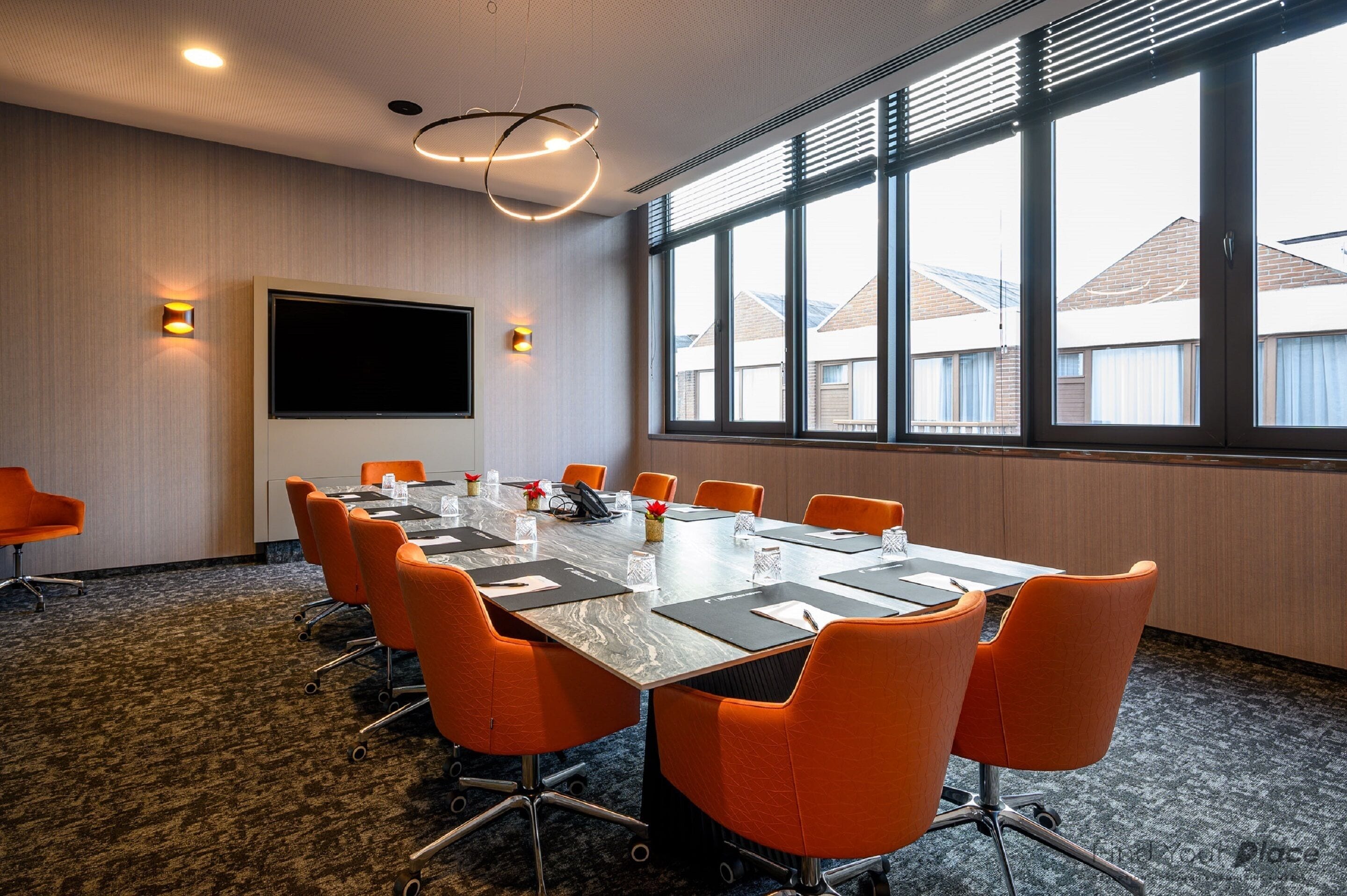 Een vergaderruimte met een grote tafel, oranje stoelen, een flatscreen televisie en uitzicht op gebouwen door grote ramen.
