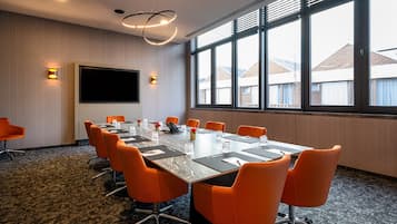 Een vergaderruimte met een grote tafel, oranje stoelen, een flatscreen televisie en uitzicht op gebouwen door grote ramen.