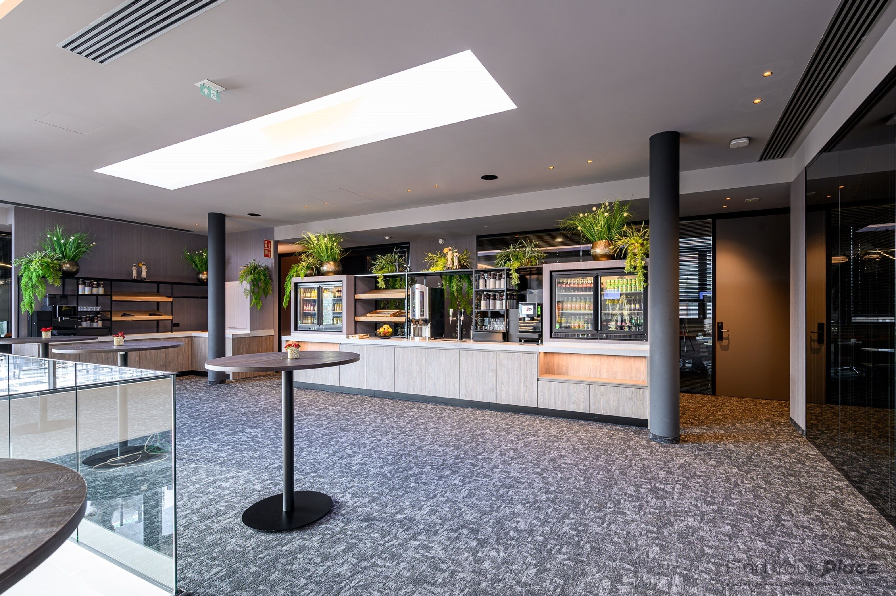 A modern lobby with a glass partition, seating area, and a bar with plants and beverages.
