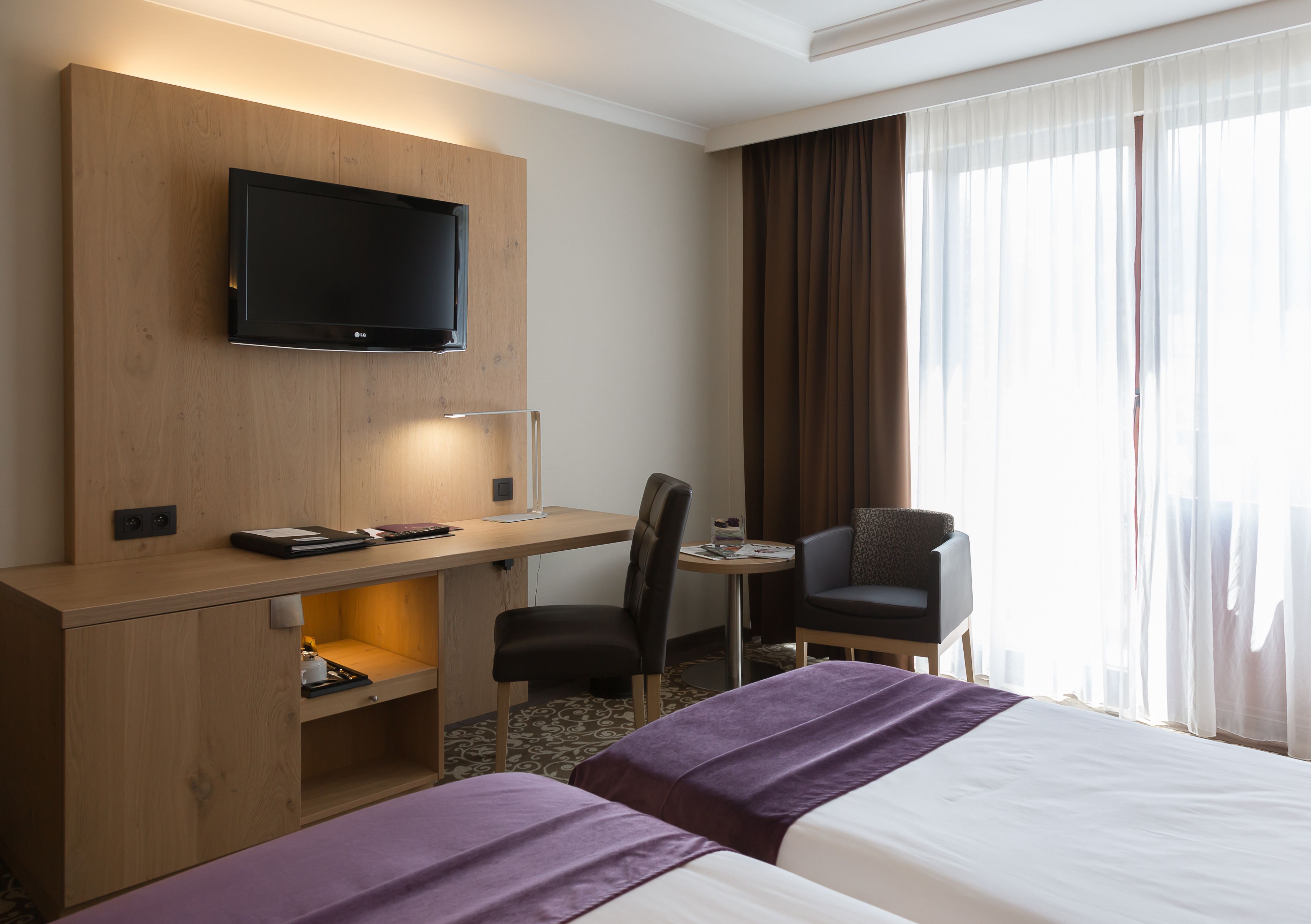 A hotel room with a bed, a desk with a chair, a television mounted on the wall, and a window with curtains.