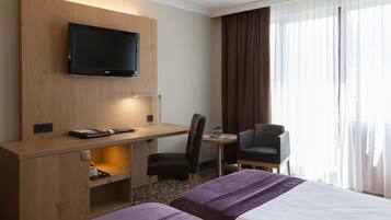 A hotel room with a bed, a desk with a chair, a television mounted on the wall, and a window with curtains.