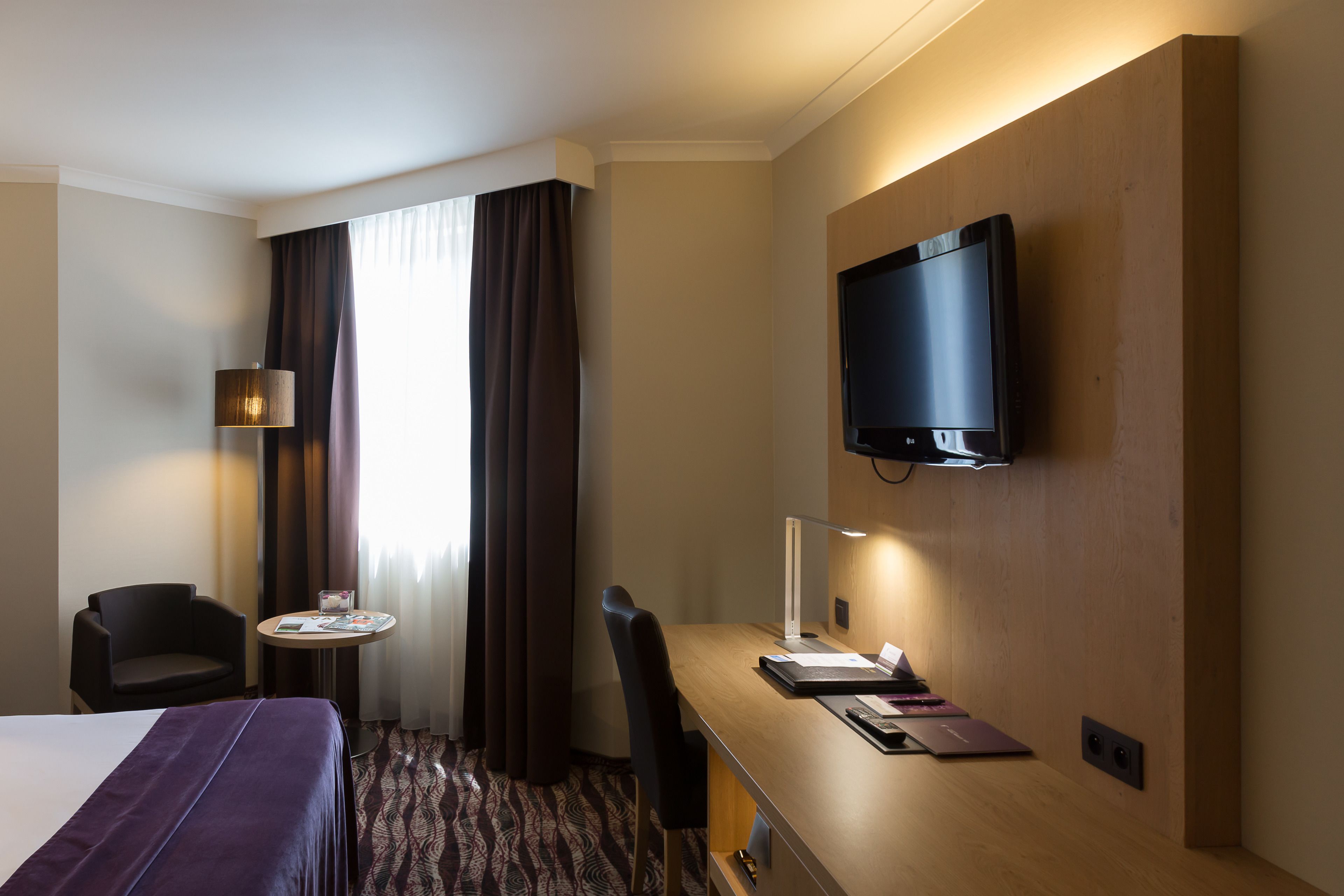 A hotel room with a bed, a desk, a chair, a television, and a small table with a lamp.