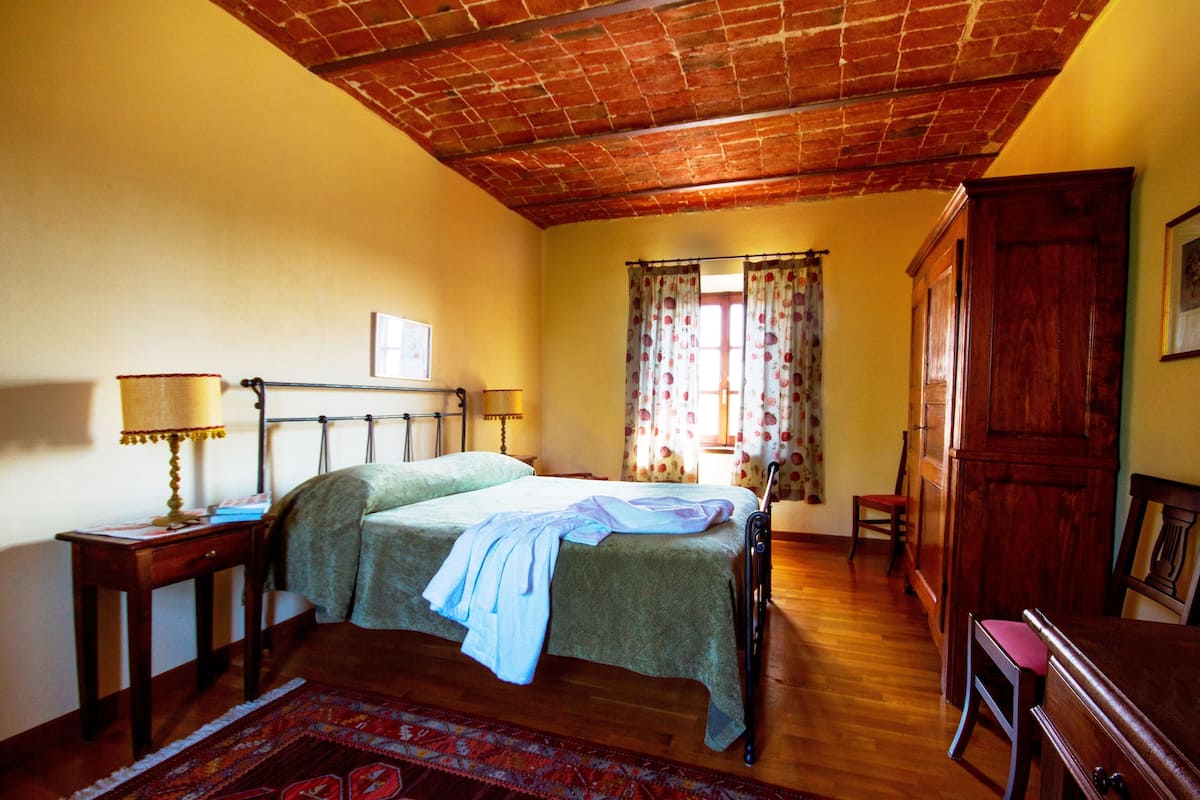A bedroom with a wooden ceiling, a bed with a green bedspread, a nightstand with a lamp, a wooden wardrobe, and a window with curtains.