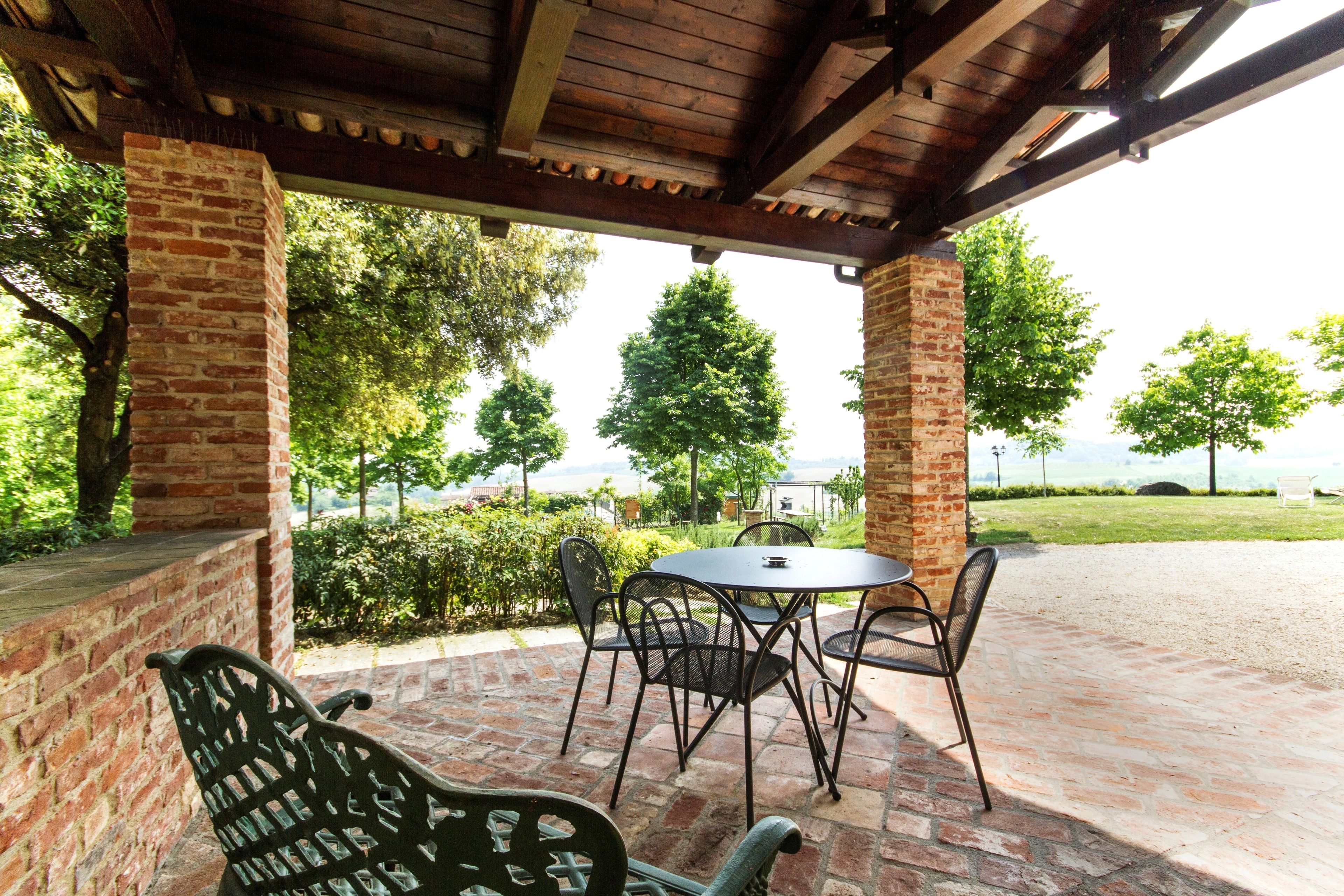 Un patio techado con pared de ladrillo, una mesa con sillas y vistas a árboles y un cuerpo de agua.