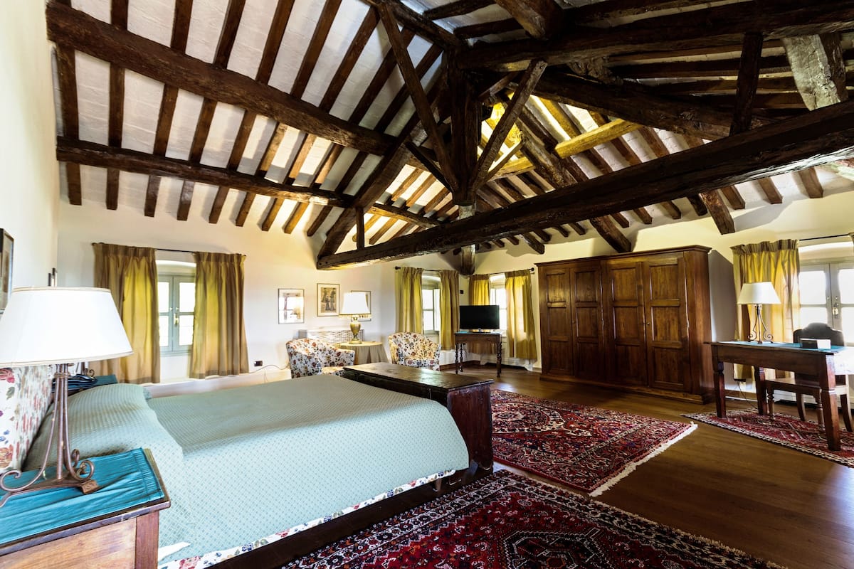 A spacious room with a large bed, wooden beams on the ceiling, a desk, and a television.