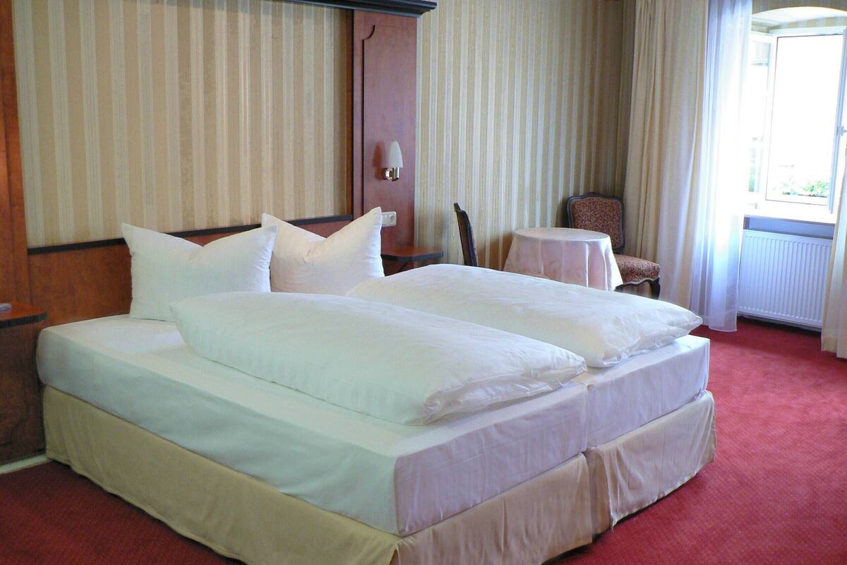 A double bed with white linens, a small round table, and a chair in a room with striped wallpaper and a red carpet.