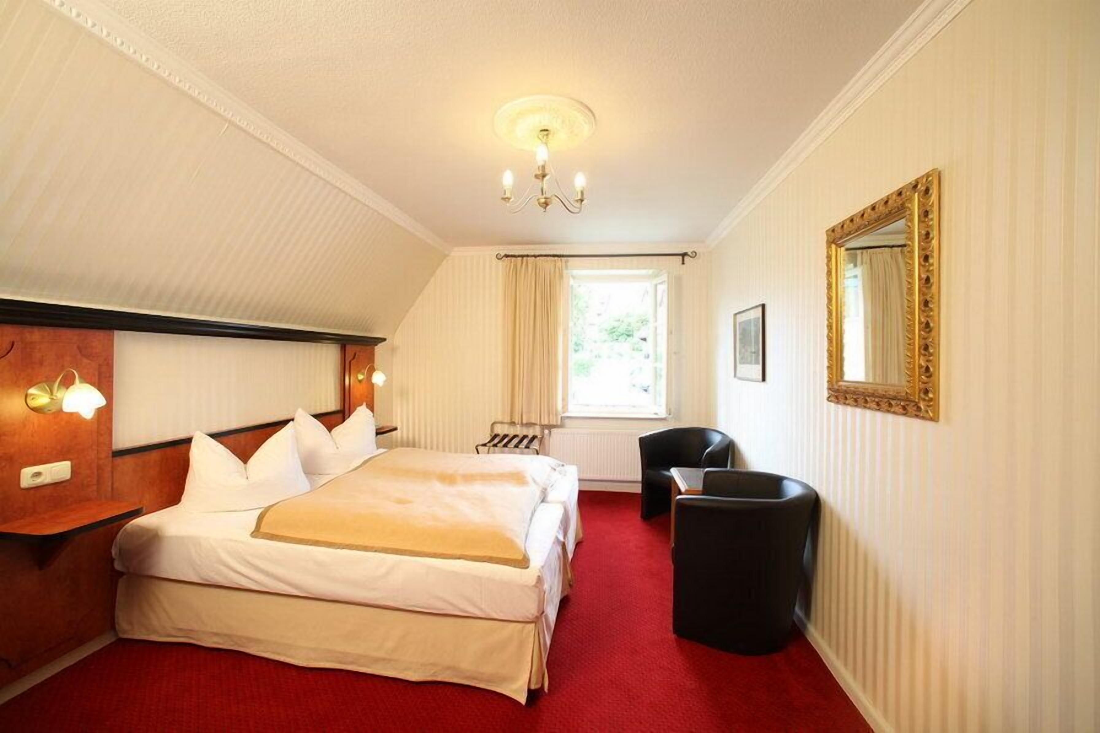 Een hotelkamer met een groot bed, een bureau, een stoel, een raam met gordijnen, een kroonluchter en een spiegel aan de muur.