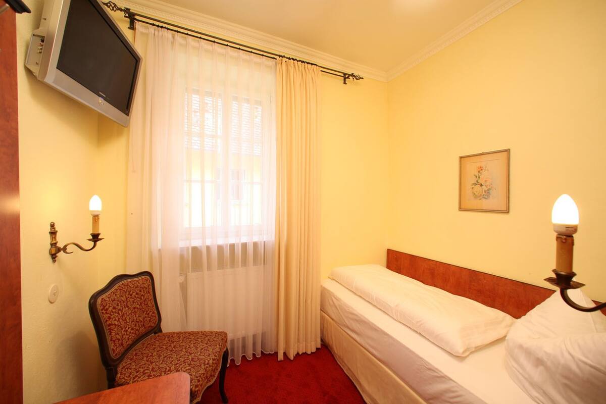 A hotel room with a single bed, a chair, a television mounted on the wall, and a window with curtains.