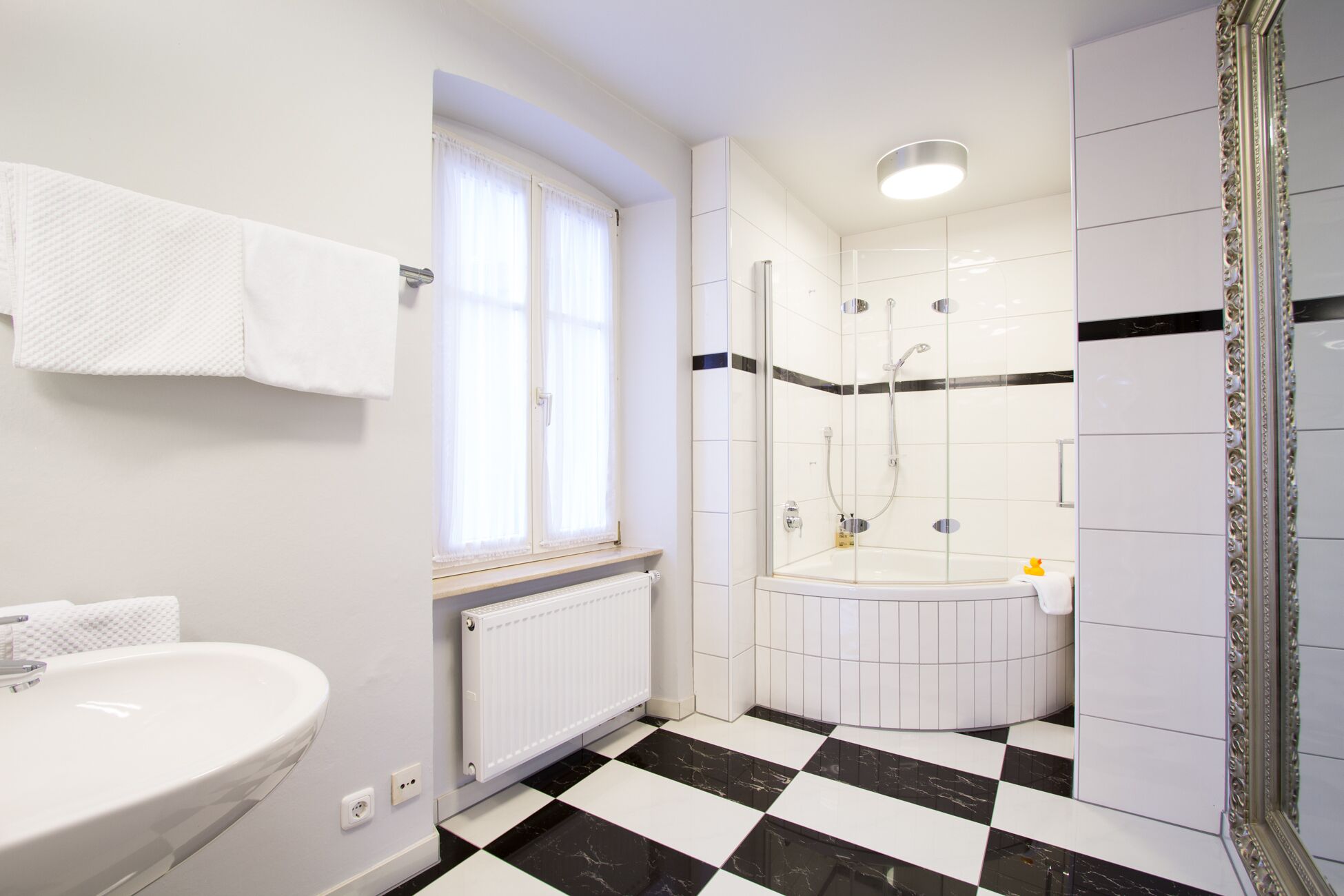 Ein Badezimmer mit einem weißen Waschbecken, einem Fenster mit einem an der Wand hängenden Handtuch, einer Dusche mit Glastür und einem karierten Boden.