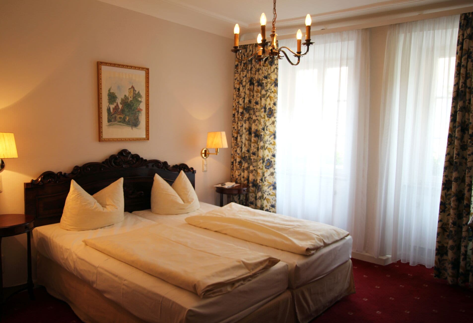 A hotel room with a large bed, a chandelier, floral curtains, and a painting on the wall.