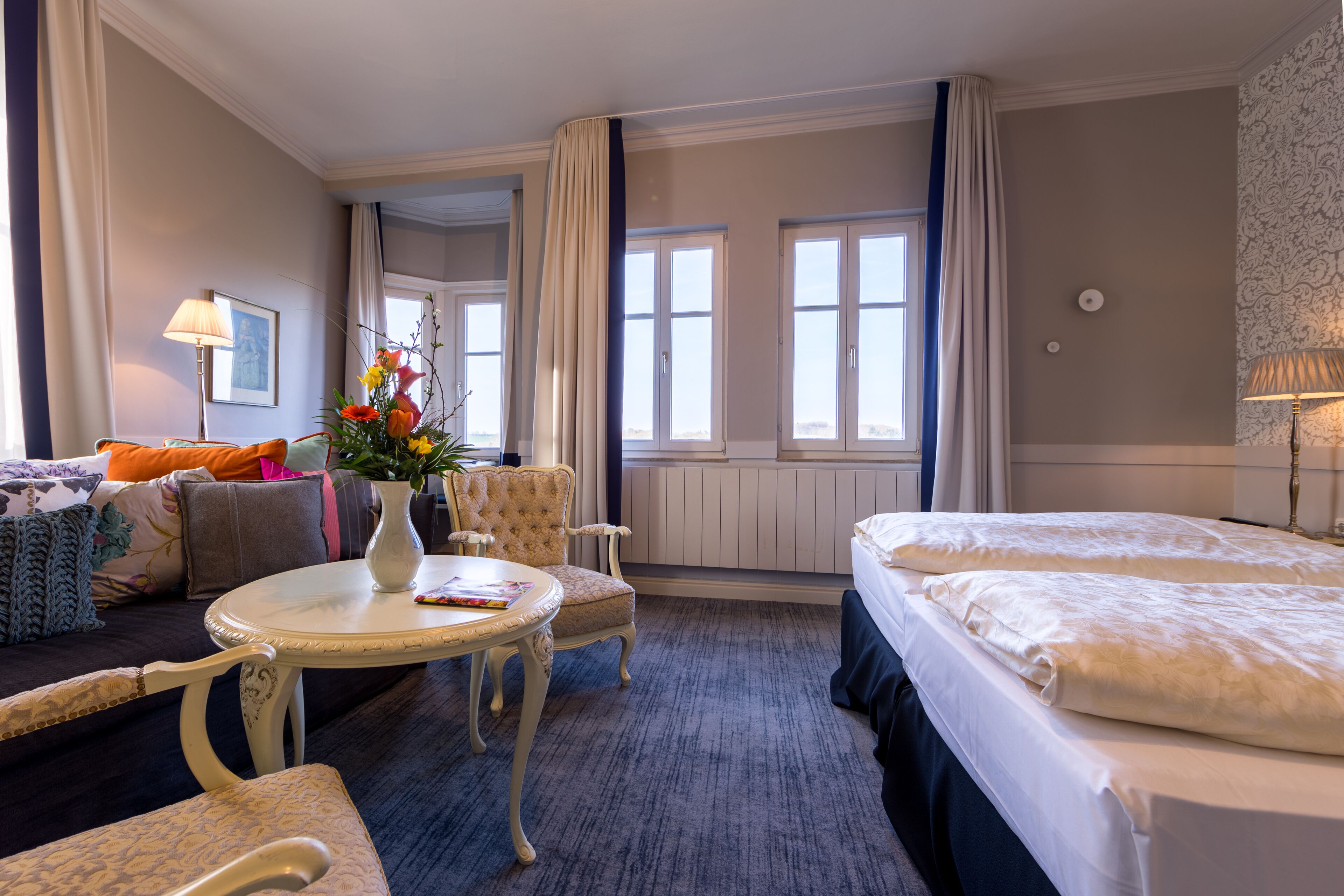 Una habitación de hotel con cama, sofá, sillón, mesita y un florero con flores.
