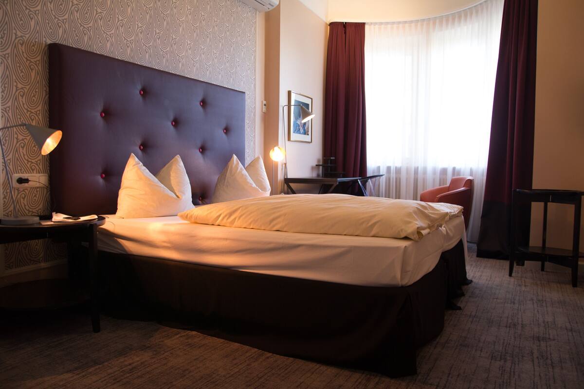 A hotel room with a large bed, a bedside table with a lamp, a small table, a chair, and a window with curtains.