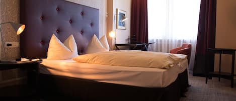 Habitación de hotel con una cama grande, una mesita de noche con lámpara, una pequeña mesa, una silla y una ventana con cortinas.
