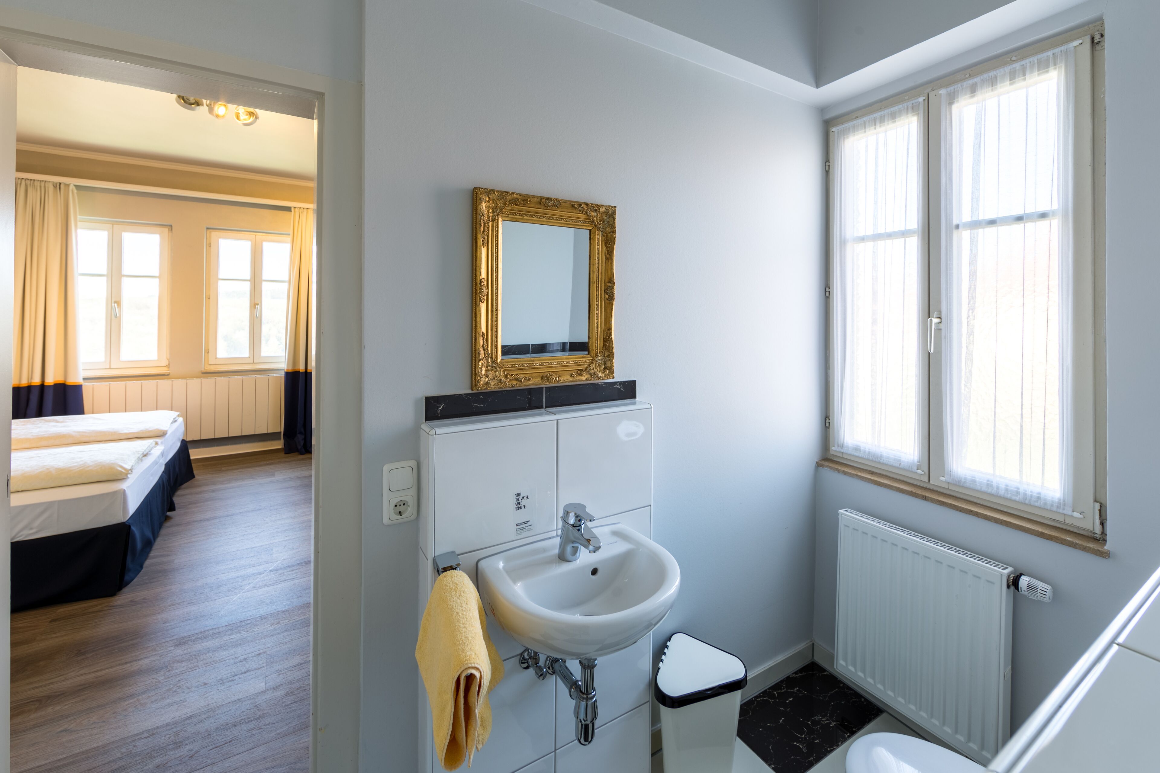 A hotel bathroom with a white sink, a gold-framed mirror, and a window with sheer curtains.