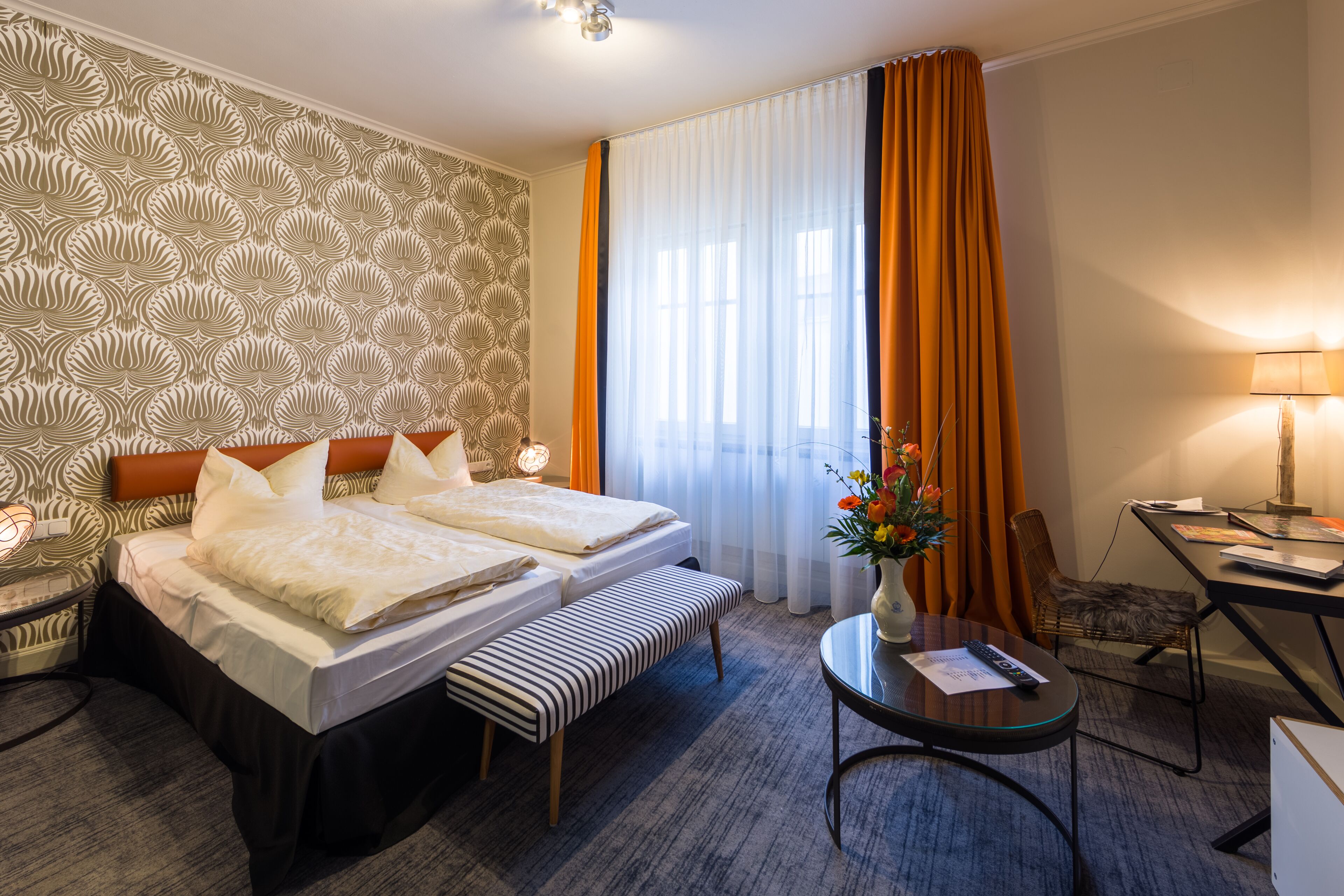 A hotel room with a double bed, a desk with a chair, a small table, a lamp, and a vase with flowers.