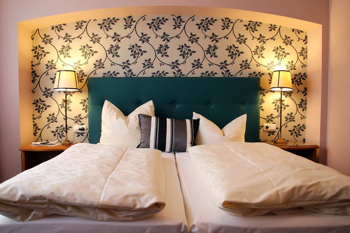 A double bed with a teal headboard, two bedside lamps, and floral wallpaper.