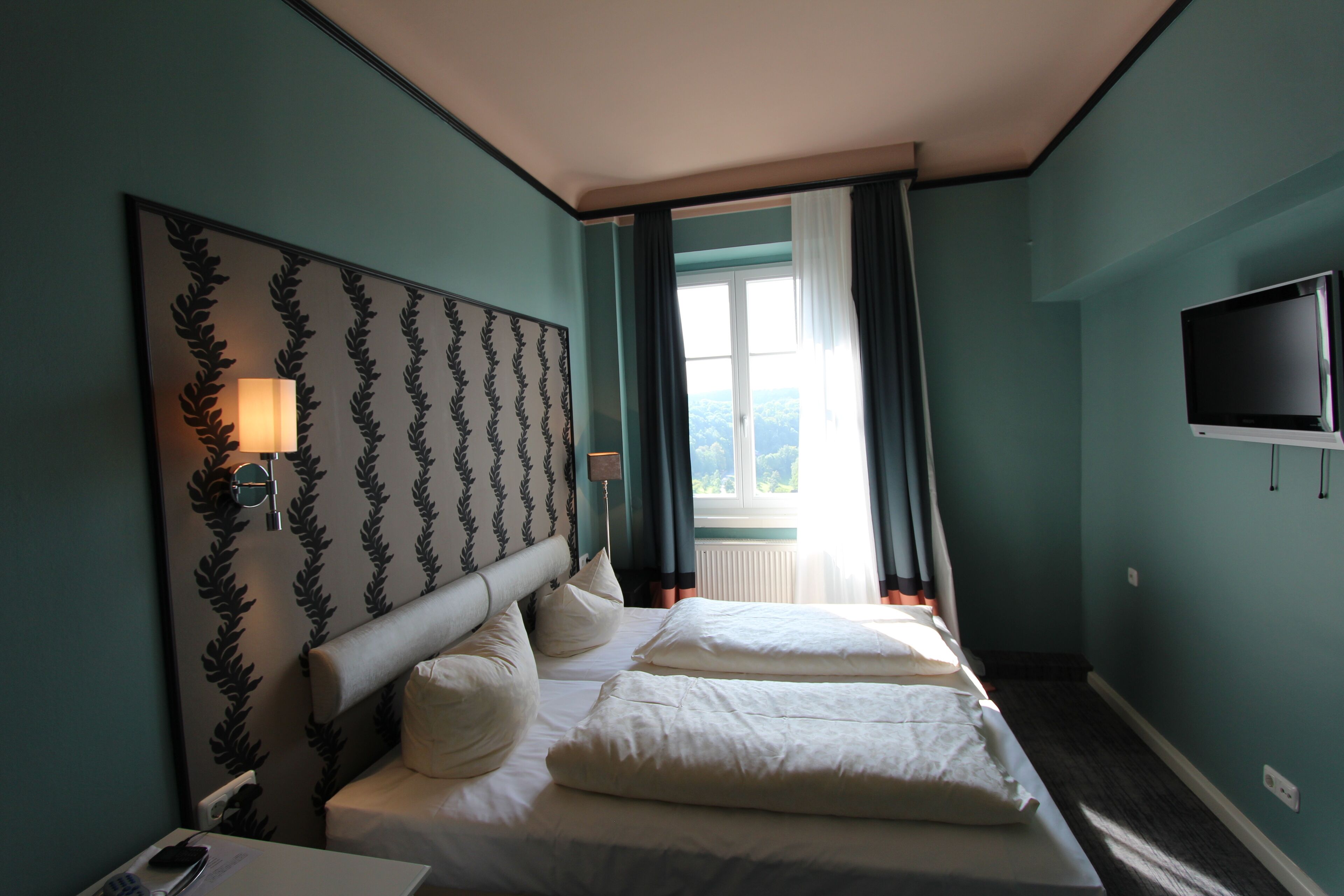 Quarto de hotel com uma cama grande, cabeceira com padrão de folha, luminária de cabeceira, televisão e janela com cortinas.