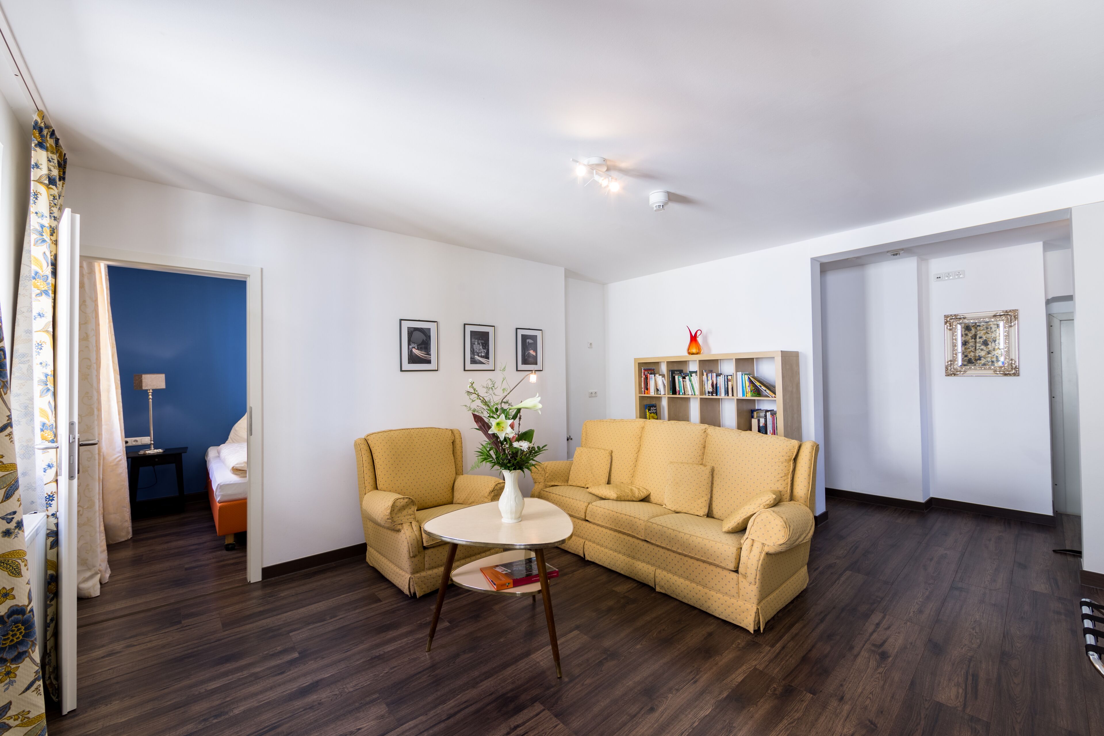 A living room with a sofa, armchair, and a small table with a vase. There is a bookshelf and a door leading to another room.