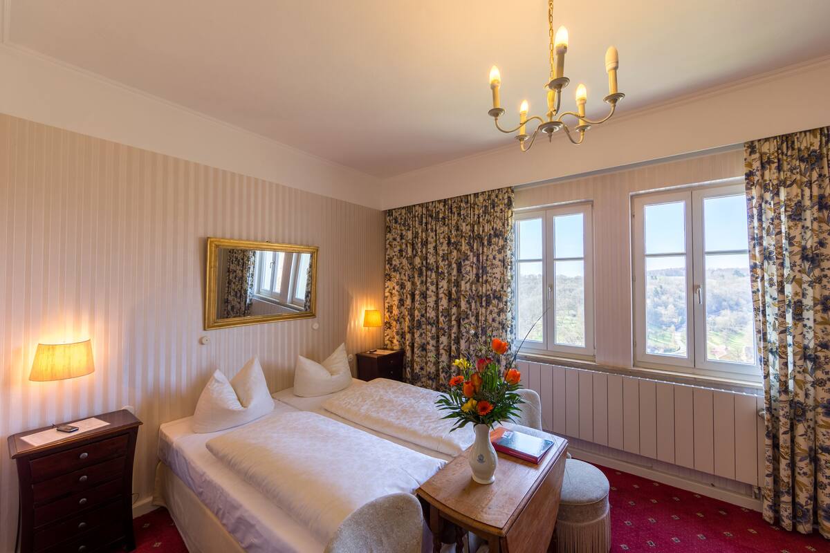 A hotel room with a large bed, bedside lamps, a mirror, a desk with a vase of flowers, and a view through the window.
