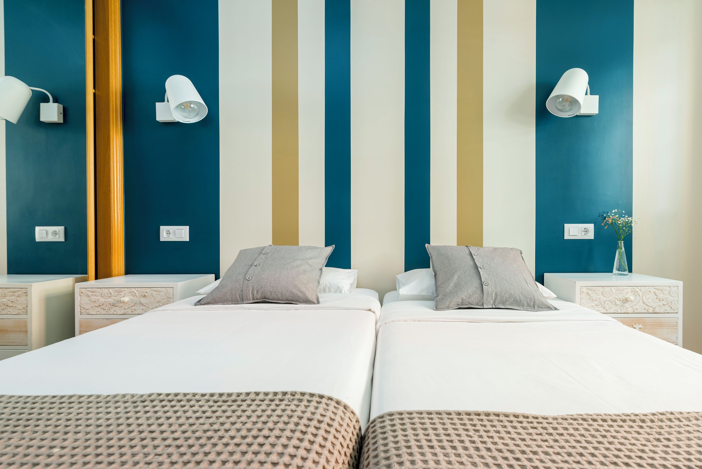 Quarto de hotel com duas camas, uma mesa de cabeceira com abajur e parede com listras verticais.
