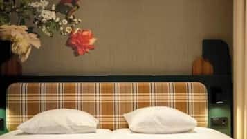 Ein Hotelzimmer mit einem Doppelbett, zwei Kissen, einem Vorhang mit Karomuster, einem Nachttisch mit Lampe und einer Blumenkomposition an der Wand.