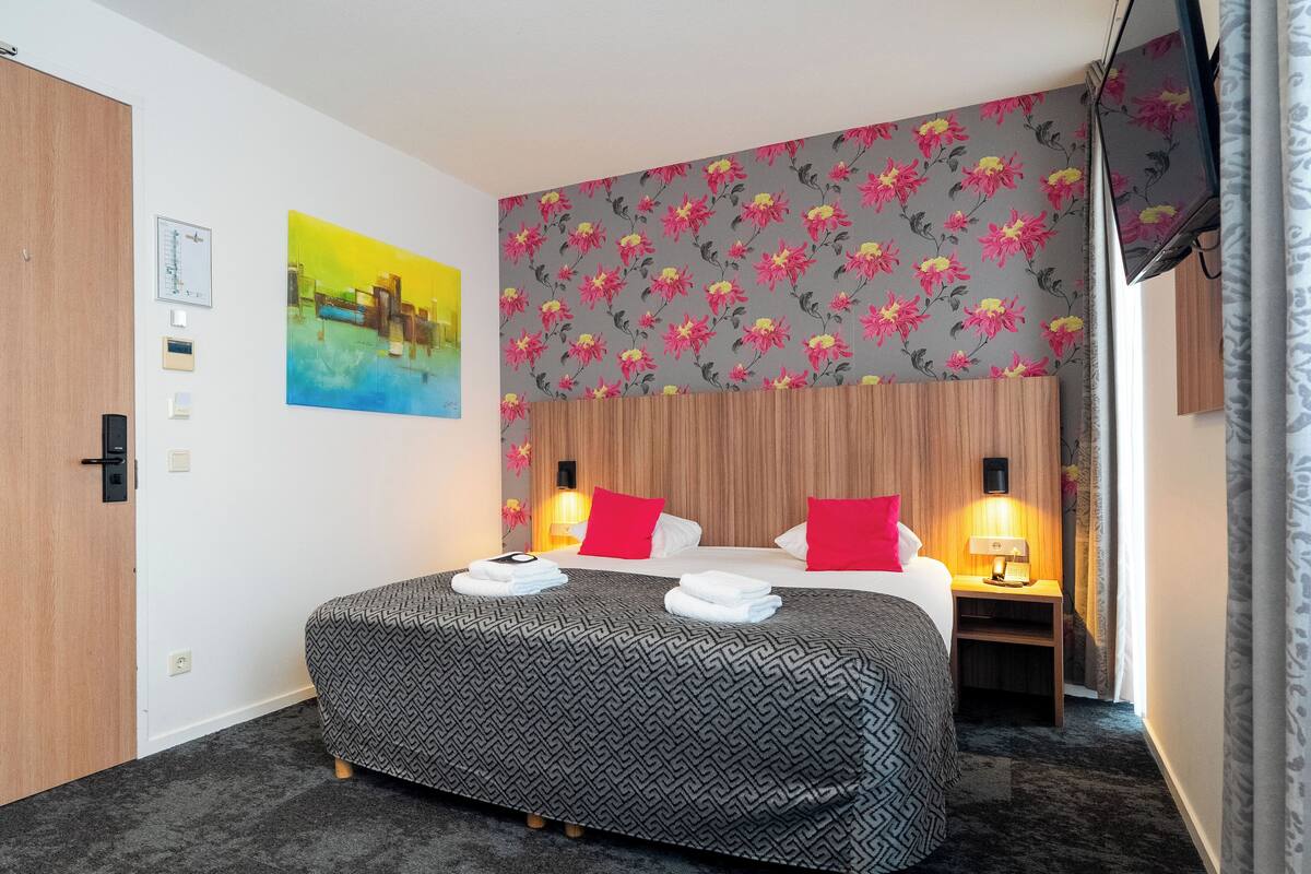 A hotel room with a bed, a nightstand, a TV, and a floral wallpaper.