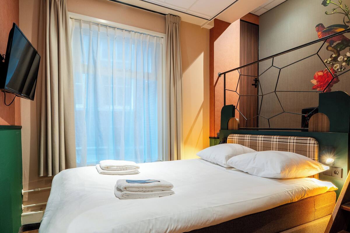 A hotel room with a bed, a TV, a window with curtains, and a decorative wall piece.
