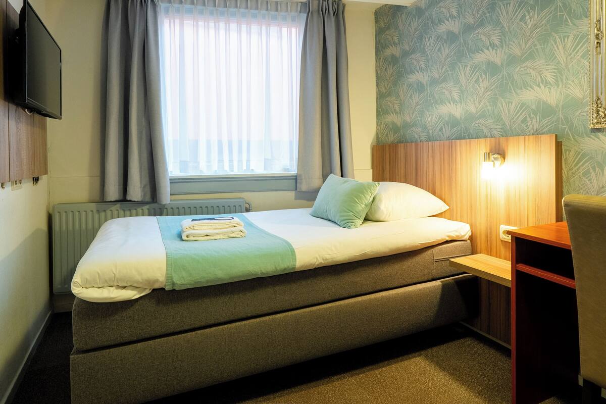A hotel room with a bed, a television, a window with curtains, a radiator, and a wall with a floral wallpaper pattern.